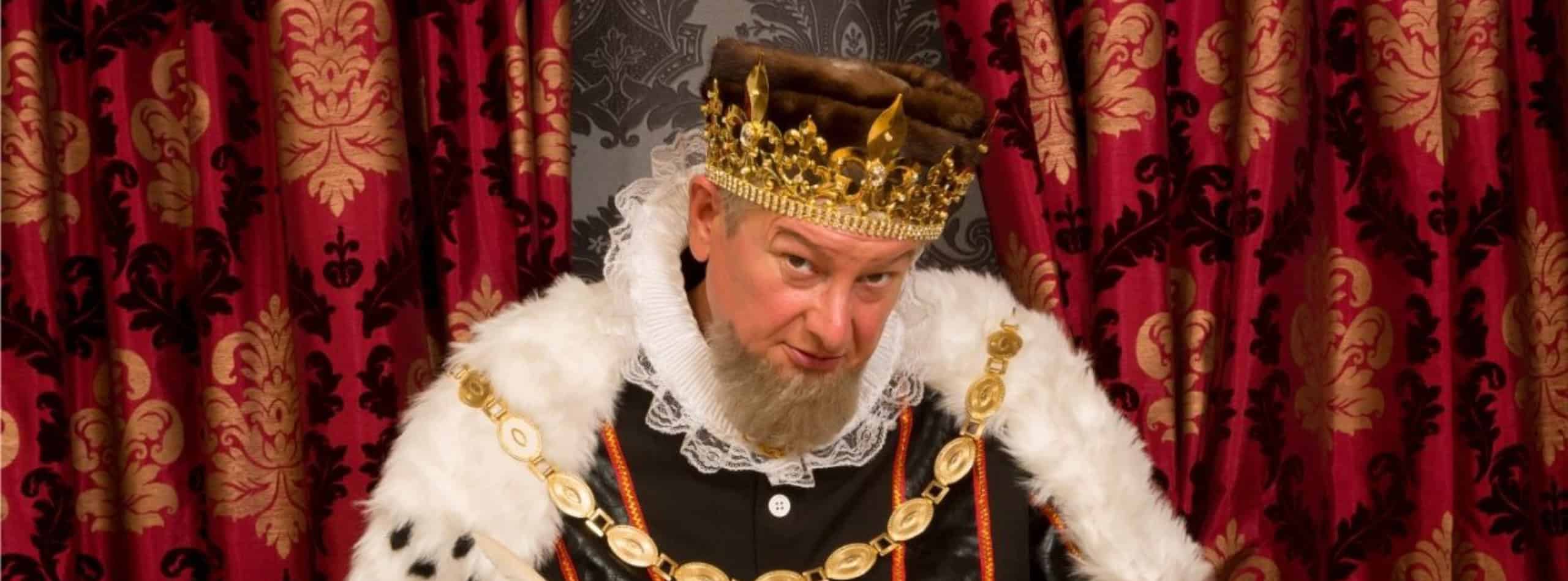 Mann in Königskrone und Hermelinmantel vor rotem Samtvorhang.